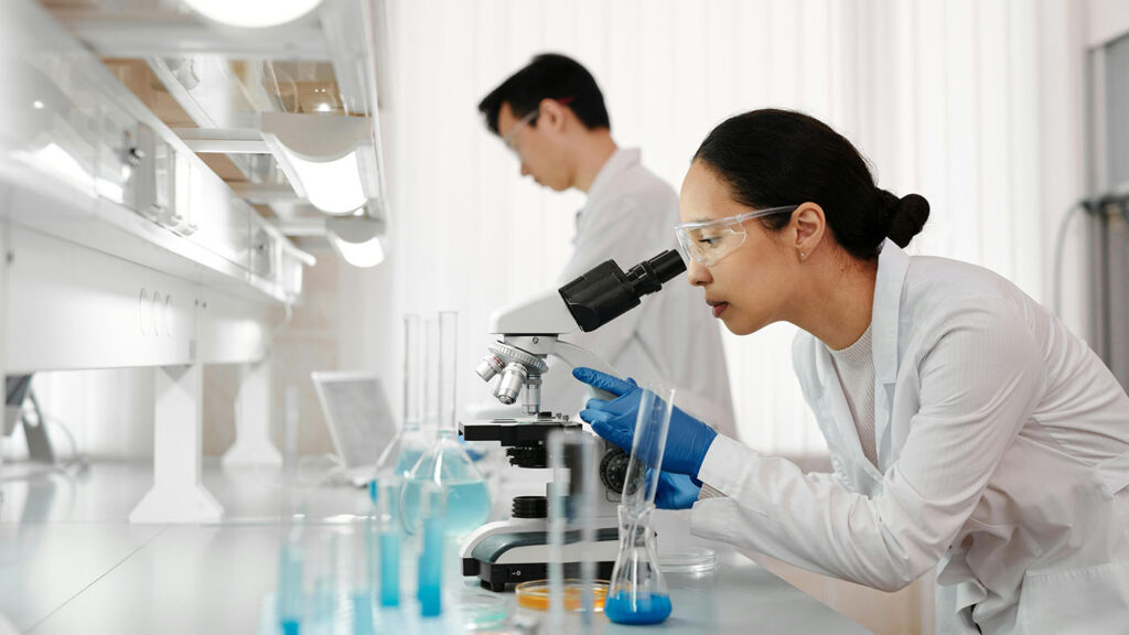 Two lab technicians working in a regulated facility, with one examining samples under a microscope as part of the Psilocybin Laboratory License process.