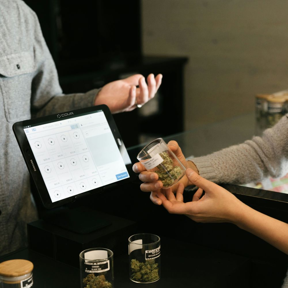 Budtender showing cannabis product to customer in front of dispensary POS touchscreen